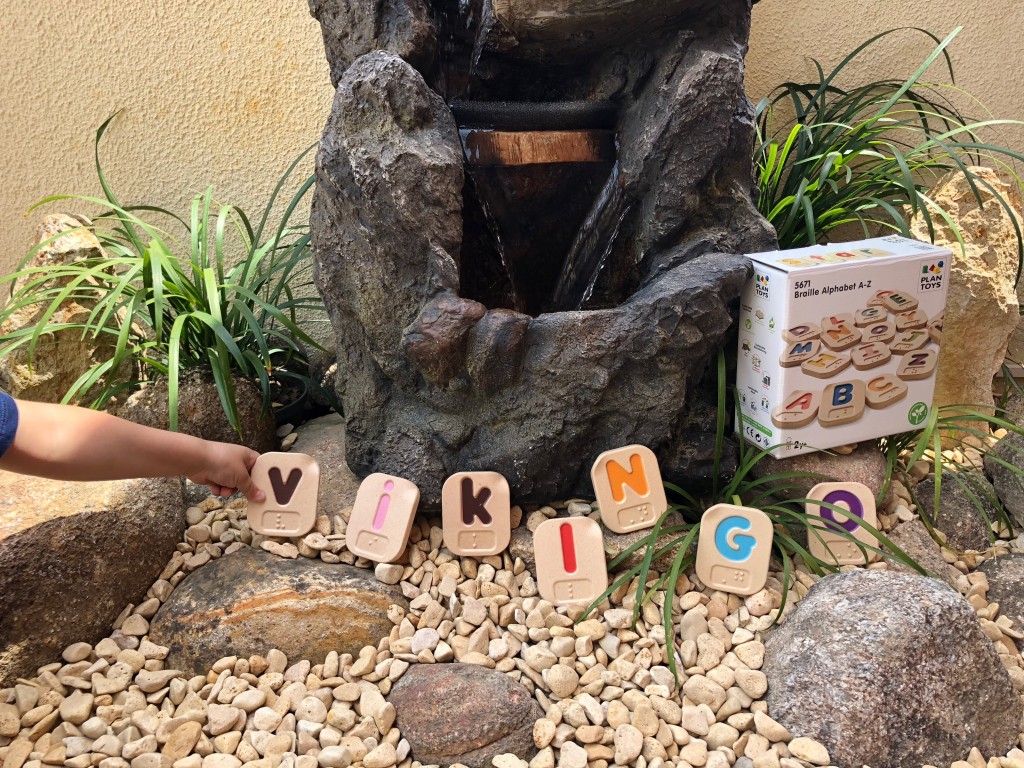 En la fuente de nuevo, entre las piedrecitas y las plantas se ve la caja del juguete, las letras formando la palabra vikingo y la mano del Vikingo sujetando la V.